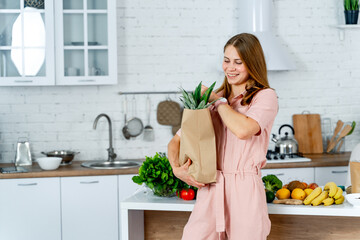 Canvas Print - A woman is holding a bag of groceries in a kitchen. She is smiling and she is happy. The kitchen is well-equipped with a sink