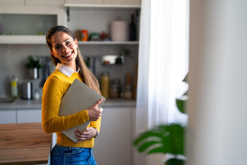 Wall Mural - Portrait photo of a smiling woman entrepreneur looking at camera while standing in a home office and holding a laptop.