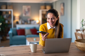 Wall Mural - Happy beautiful businesswoman messaging online on smart phone while using laptop at home.