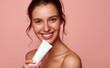 © Curioso.Photography - A smiling woman holding a tube of skincare product against a pink background.