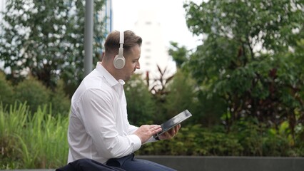 Wall Mural - Businessman wear his headphone to listen relaxed music while take off suit and move to music at green city. Manager with headset dancing movement with lively song while hold phone. Back view. Urbane.