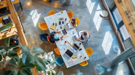 The photo shows a group of people sitting around a table in an office space. They are all working on different projects and using different devices. The space is modern and well-lit, with a variety of