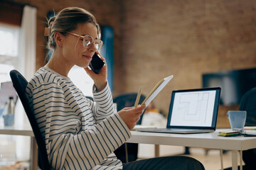 Young female architect is talking phone with colleague and looking on notepad sitting in coworking