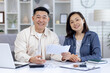 © Liubomir - Happy asian couple sitting at table managing their finances using a laptop and documents, showing a positive family bond.