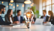 © Your Hand Please - glowing light bulb on a white tabletop symbolizes innovation and bright ideas, with blurred business people meeting in the background, emphasizing collaboration and creativity