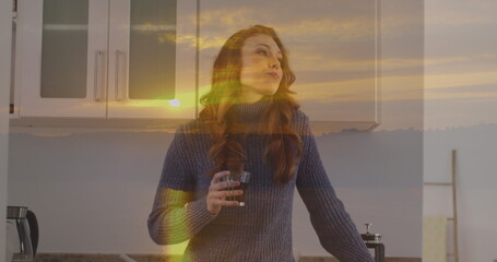 Image of sunset landscape over caucasian woman drinking coffee in kitchen