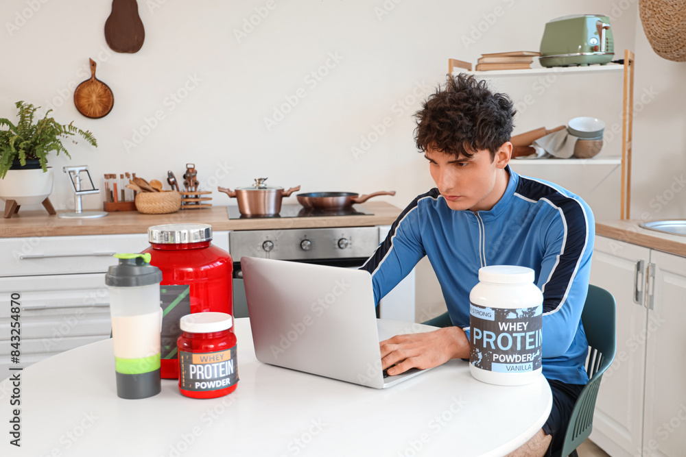 Sporty young man with protein powder using laptop at table in kitchen
