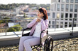 © sofiko14 - Woman in wheelchair enjoying the view of the city from a rooftop on a sunny day. She appears relaxed and content, wearing a casual outfit. Urban landscape and buildings in the background.
