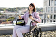 © sofiko14 - Woman in wheelchair on rooftop holding coffee cup and smartphone, enjoying sunlight. Urban cityscape in background, demonstrating independence and technology use.