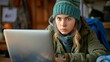 © Sergey - A focused young woman in winter attire working on a laptop in a cozy indoor setting.