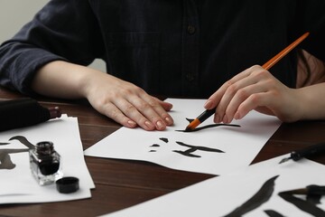 Wall Mural - Calligraphy. Woman with brush and inkwell writing hieroglyphs on paper at wooden table, closeup