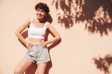 Young beautiful smiling hipster woman in trendy summer shorts and top white t-shirt. Carefree woman posing in the street near wall at sunset. Positive model outdoors. Cheerful and happy