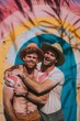 © Glittering Humanity - Two men embracing in front of a rainbow painted wall, showing affection and pride