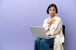 © luismolinero - Young mixed race woman sitting on a chair with laptop isolated on purple background pointing to oneself