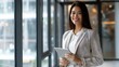 © The Little Hut - Young happy businesswoman using digital tablet while standing by the window in the office and looking at camera Copy space : Generative AI