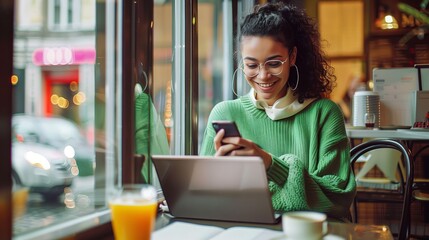 Wall Mural - A woman sitting at the window of an open coffee shop, holding her phone and smiling as she reads something on social media while sipping tea from one hand with a laptop in front of her