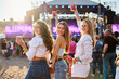 © artiemedvedev - Women at beachfront music fest enjoy drinks, dance, smile in sun. Girl group holds cocktails, celebrates summer event. Carefree moments, lively festival atmosphere with stage in background.