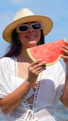 Wall Mural - A woman is standing on a beach holding a watermelon slice. She is wearing a white bikini and a straw hat. The beach is rocky and the sky is clear