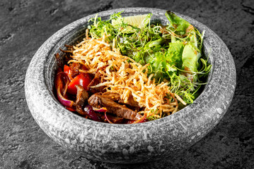 Wall Mural - Stone bowl of salad with greens, shredded vegetables, meat strips, and crispy onions on a dark textured surface. Ideal for culinary and healthy eating concepts