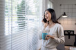 © amnaj - Woman talking on phone and holding coffee cup while standing by window with blinds in modern home office, bright and relaxed atmosphere.