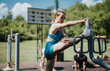 © qunica.com - Young woman in athletic wear performing a leg stretch and exercise outdoors at a fitness park. Sunny day, green trees, focused on fitness and flexibility.