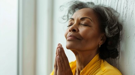 Wall Mural - A woman with closed eyes wearing a yellow top has her hands clasped together in a prayerful pose leaning against a white wall with a window in the background.