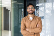 © Tetiana - Portrait of a young Indian man wearing glasses and a brown shirt standing in a modern office, crossing his arms over his chest and looking confidently at the camera