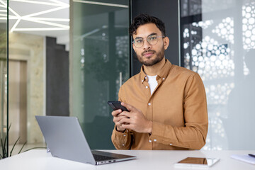 Wall Mural - Portrait of a young Indian man in a brown shirt and glasses sitting at a desk in the office, holding a phone and looking at the camera