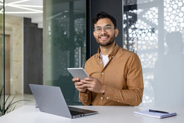 Wall Mural - Portrait of a young Muslim man wearing glasses sitting at an office desk with a laptop, holding and using a tablet, looking at the camera with a smile