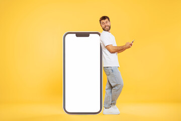 Cheerful European bearded man using cellphone while standing near huge smartphone with empty screen on yellow studio background, mock up for website app for advert