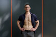 © arthurhidden - A self-assured man standing confidently in an indoor setting with his shirt half-open, showcasing his fit physique. The neutral background accentuates his composed demeanor and casual style.