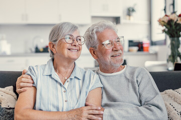 Canvas Print - Dreamy middle aged senior loving retired family couple looking in distance, planning common future or recollecting memories, enjoying peaceful moment relaxing together on cozy sofa in living room.