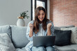 © Daniel - Excited emotional screams with joy woman sit on sofa hold on lap notebook makes Yes hand gesture feels happy getting job of dream, e-mail sales discount special offer, on-line lottery victory concept.