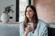 © Daniel - Smiling woman looking at smartphone screen, sitting on couch, enjoying leisure time with mobile device at home, happy young female chatting in social network or surfing internet, shopping online.