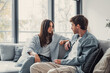 © Daniel - Young happy couple having fun talking laughing relaxing at home on couch, boyfriend embracing girlfriend telling funny joke sitting on sofa, humor in relationships, enjoying weekend together.
