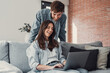 © Daniel - Happy millennial couple sit relax on couch in living room, smiling young husband standing from back and wife rest on sofa at home browsing Internet using modern computer device.