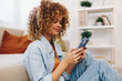 © SHOTPRIME STUDIO - Cozy Living: Young Woman Holding Phone and Smiling while Relaxing on Sofa in her Home