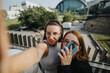 © qunica.com - Two friends enjoying their time together outdoors, capturing a fun selfie on a sunny day with a modern urban background.