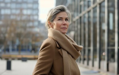 A woman wearing a brown coat and scarf stands in front of a building. She has a serious expression on her face