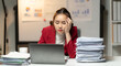 © Nittaya - Overwhelmed Businesswoman:  A young Asian businesswoman sits at her desk, looking stressed and tired, surrounded by a large stack of paperwork and a laptop.