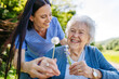 © Halfpoint - Female caregiver and senior woman in wheelchair holding dandelion, picking wild flowers. Nurse and elderly woman enjoying a warm day in nursing home, public park.