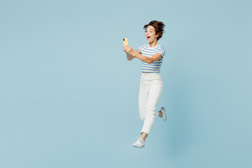 Full body side view young woman wear striped t-shirt casual clothes jump high hold in hand use mobile cell phone isolated on plain pastel light blue cyan background studio portrait. Lifestyle concept