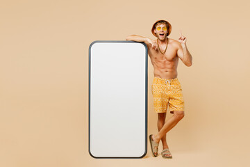 Full body young man wears yellow shorts swimsuit hat relax rest near hotel pool big blank screen mobile cell phone point finger up isolated on plain beige background. Summer hot sea sun tan concept.