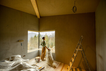 Young woman stands with her dog and looks out of the window while making repairment of a new house on nature. Creative process of home renovation and repair, friendship with pets concept