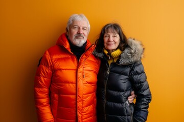 Wall Mural - Portrait of a blissful couple in their 50s donning a durable down jacket while standing against soft orange background