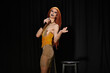 © Bliss - A drag performer in a shimmering gold dress takes the stage, microphone in hand.