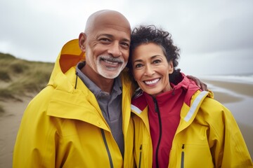 Sticker - Portrait of a happy mixed race couple in their 60s wearing a vibrant raincoat isolated on serene dune landscape background