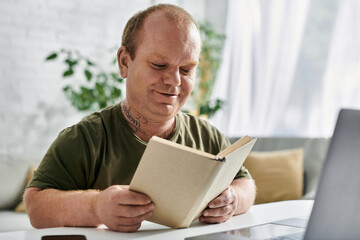 Wall Mural - A man with inclusivity sits at a table in his home, engrossed in a book. He is casually dressed in a green shirt.