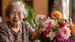 © Matcha_09 - Elderly woman with a bright, happy smile, sitting at a table with a vase of flowers.