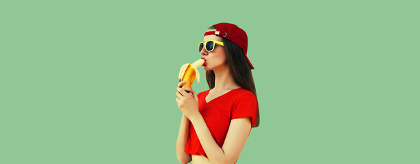 Wall Mural - Stylish young woman eating banana in red baseball cap on green studio background